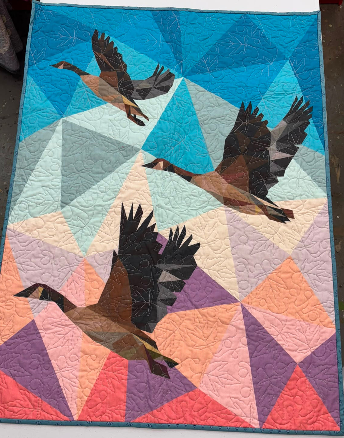 A quilted wholecloth quilt featuring three flying geese over a geometric triangle background in shades of blue, purple, pink, orange, and cream.